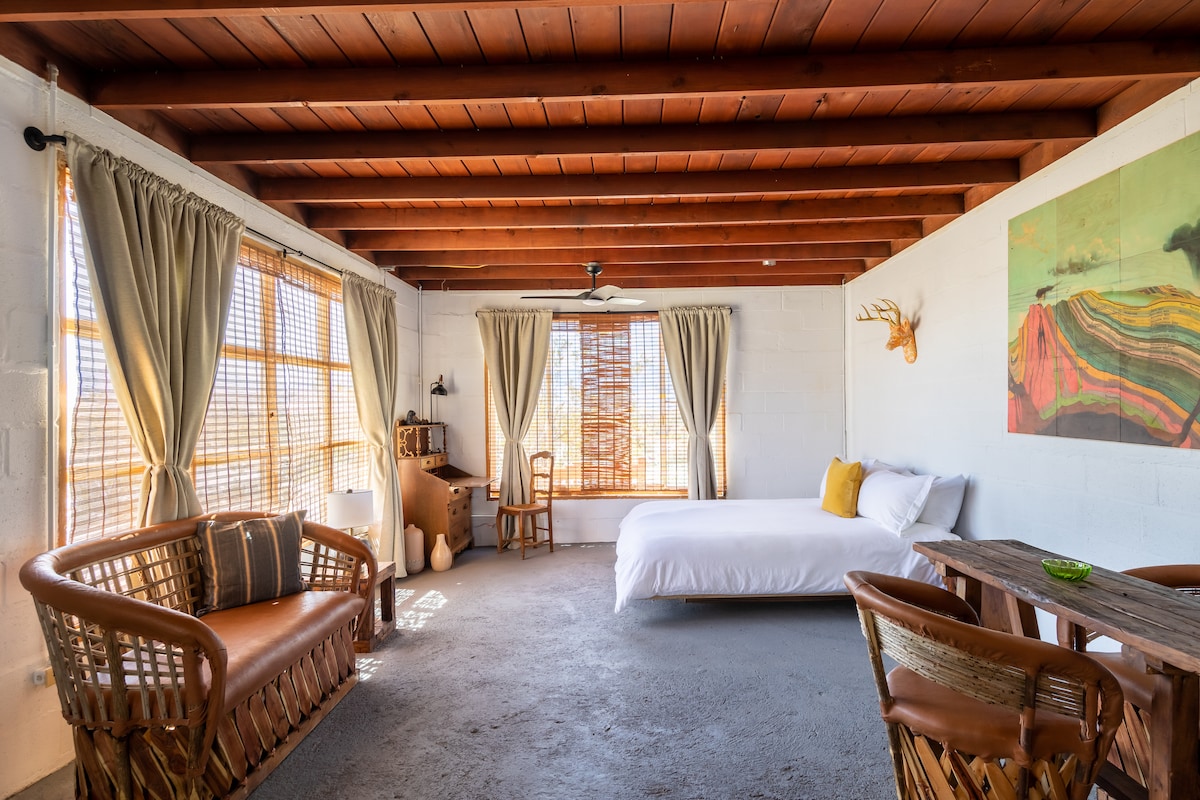 A spacious interior showcases rustic charm with wooden ceiling beams and textured concrete walls. A large bed is positioned beside a window, adorned with soft linens and decorative pillows. A seating area features woven furniture, while natural light filters through bamboo shades, enhancing the calming ambiance.