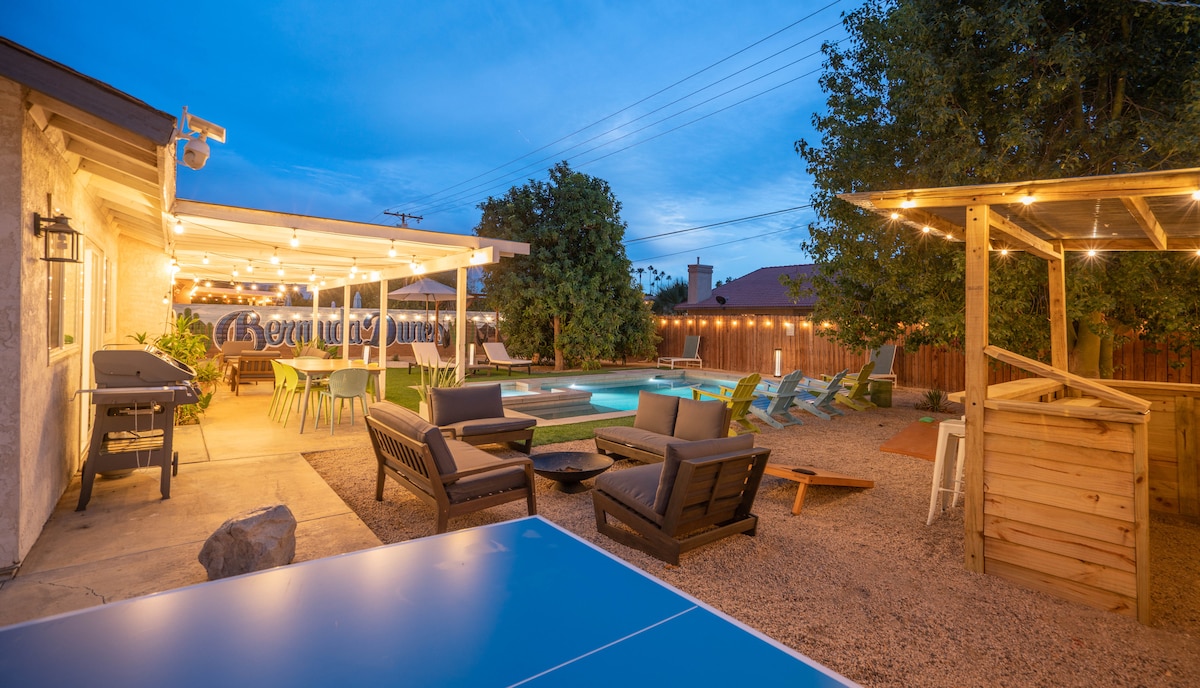 An expansive backyard is illuminated with resort-style lights, featuring a new heated pool and hot tub. Outdoor seating includes lounge chairs and a covered dining area for 8, alongside a game area with a ping pong table and a spacious hammock.