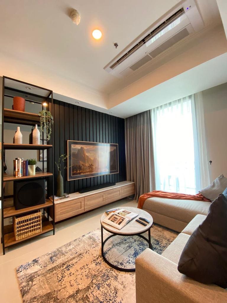 A cozy living area is featured with a light-colored sofa and an accent chair. A black and wood shelving unit displays decorative items and books. Natural light enters through large curtains, highlighting a framed artwork on the wall. A round coffee table sits in the center, complemented by a textured rug.