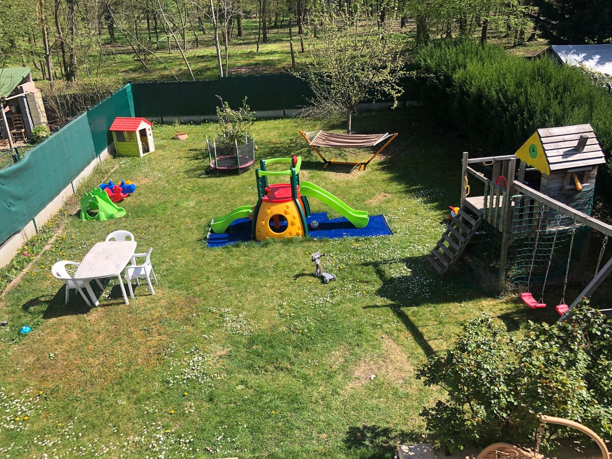 The image showcases a spacious backyard with various play equipment, including a slide, swing set, and trampoline. A small playhouse and a hammock are visible among the greenery. Garden furniture, including a table and chairs, is arranged on the lawn, surrounded by lush grass and trees.