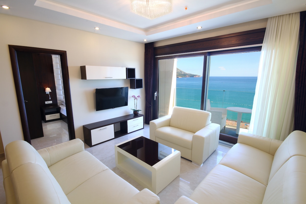 A modern living area showcases a sleek design with light-colored leather sofas arranged around a glass table. Large windows frame a panoramic view of the sea, while natural light enhances the spaciousness of the room. A flat-screen television is mounted on the wall, complemented by minimalist storage.