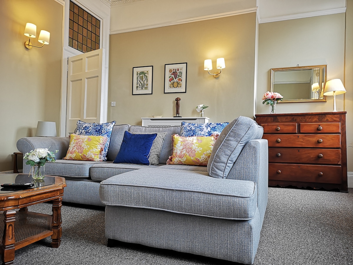 An inviting living area features a spacious grey sofa adorned with decorative cushions. A wooden coffee table displays fresh flowers, and a wooden dresser stands against the wall. Soft lighting from wall sconces adds warmth to the room, complemented by carefully placed wall art.