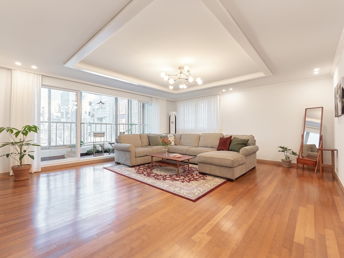 A spacious living area features a light grey sectional sofa arranged around a decorative area rug. Large windows provide ample natural light, showcasing the city view. Potted plants and modern lighting add character to the warm wooden floors and neutral-toned walls.