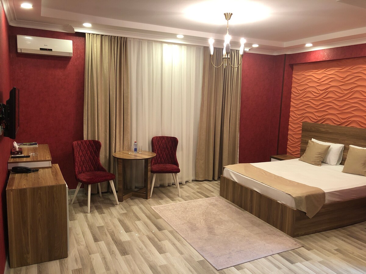 A well-furnished room features a spacious bed with neutral bedding against a textured accent wall. Two red chairs are positioned around a small table, and light-colored curtains frame a large window. A cozy area rug lies on the wooden floor, contributing to a warm ambiance.