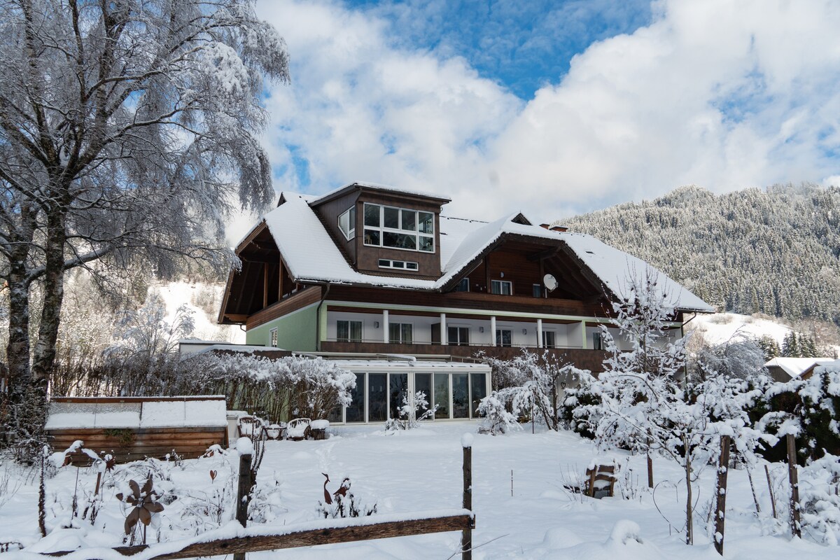 The exterior of a three-story chalet is surrounded by snow-covered trees and a tranquil winter landscape. The balcony features large glass windows, reflecting the blue sky and fluffy white clouds above, enhancing the overall wintry scene.
