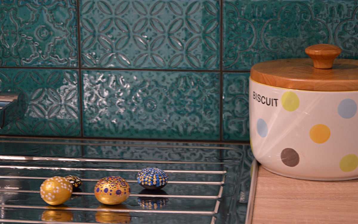 A kitchen countertop features a silver rack with decorative eggs in various colors, positioned in front of a vibrant teal backsplash. A ceramic biscuit container with a wooden lid is displayed nearby, adding a practical yet stylish element to the space.