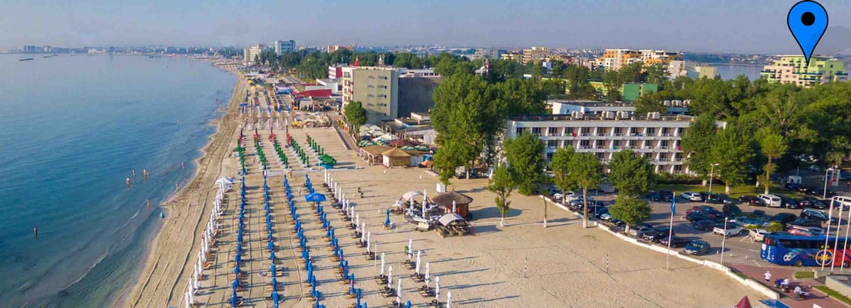 A wide view of the beach features rows of umbrellas and sun loungers arranged on the sandy shore. The clear blue water gently laps at the beach, while buildings and greenery are visible in the background, highlighting the vibrant atmosphere of the Mamaia Resort area.