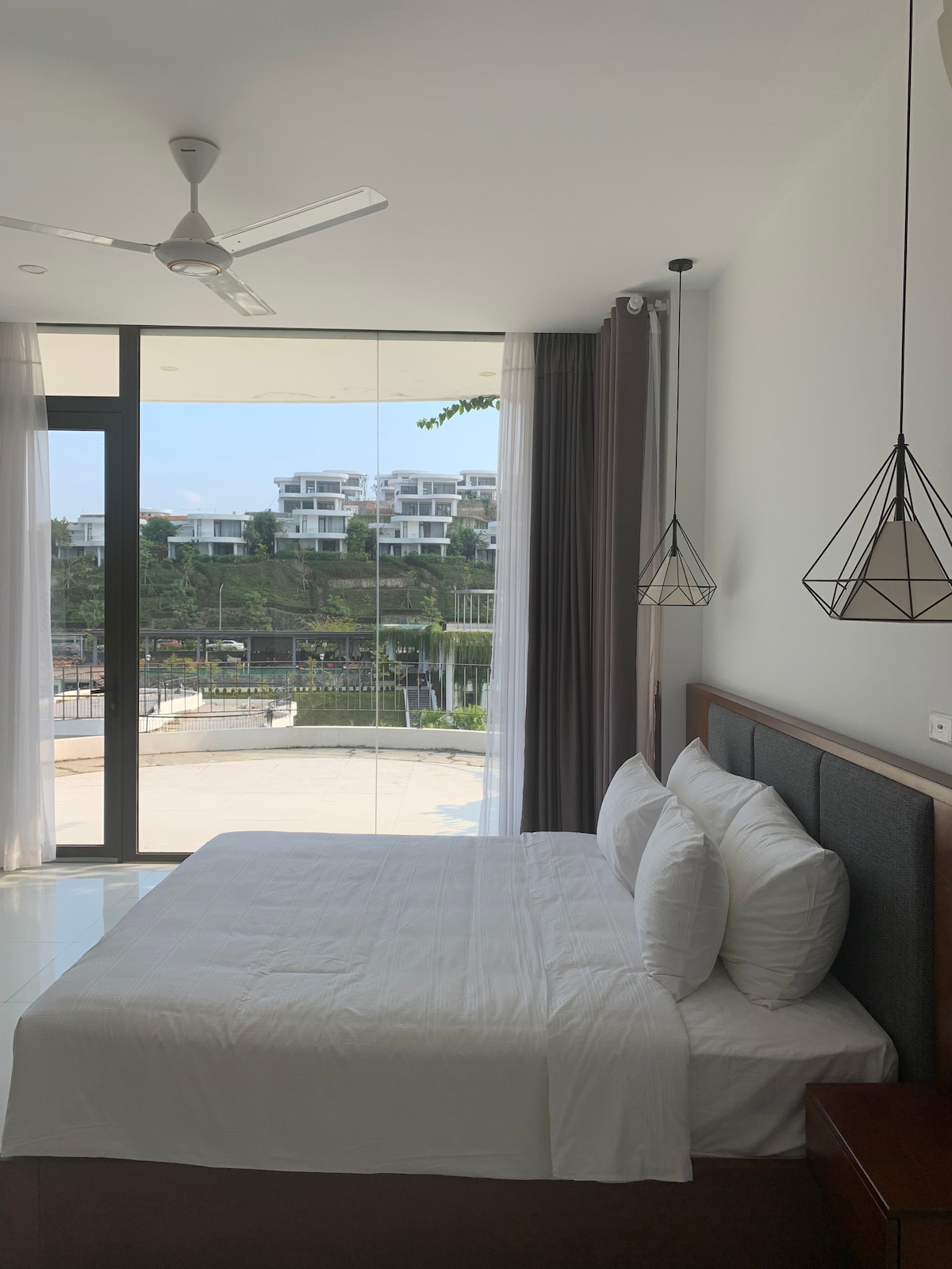 A bright bedroom features a spacious bed with multiple pillows, surrounded by large glass windows. Natural light fills the room, highlighting the clean design. Two modern pendant lights hang above, adding a subtle decorative element. Views of the resort's exterior are visible through the windows.