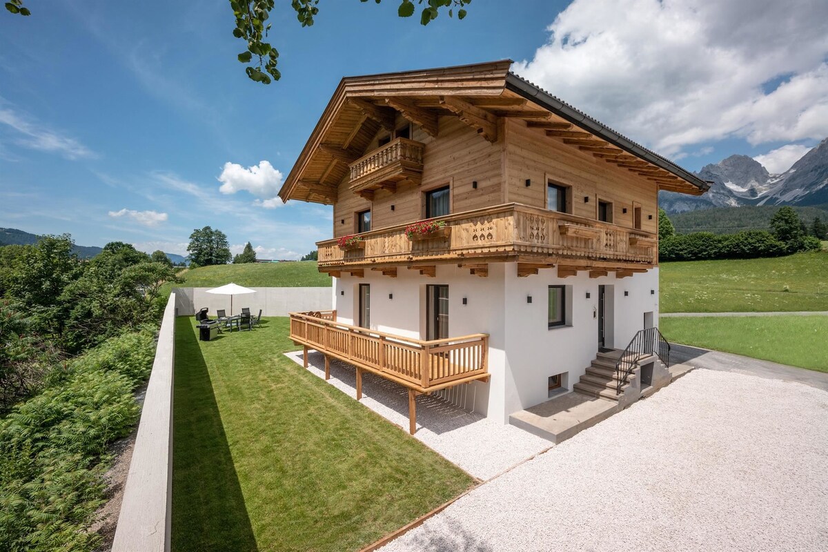 A three-story chalet exhibits traditional wooden architecture with multiple balconies. The exterior features a mix of wooden and white walls, surrounded by a manicured lawn. A seating area is visible in the foreground, with mountains visible in the background under a partly cloudy sky.