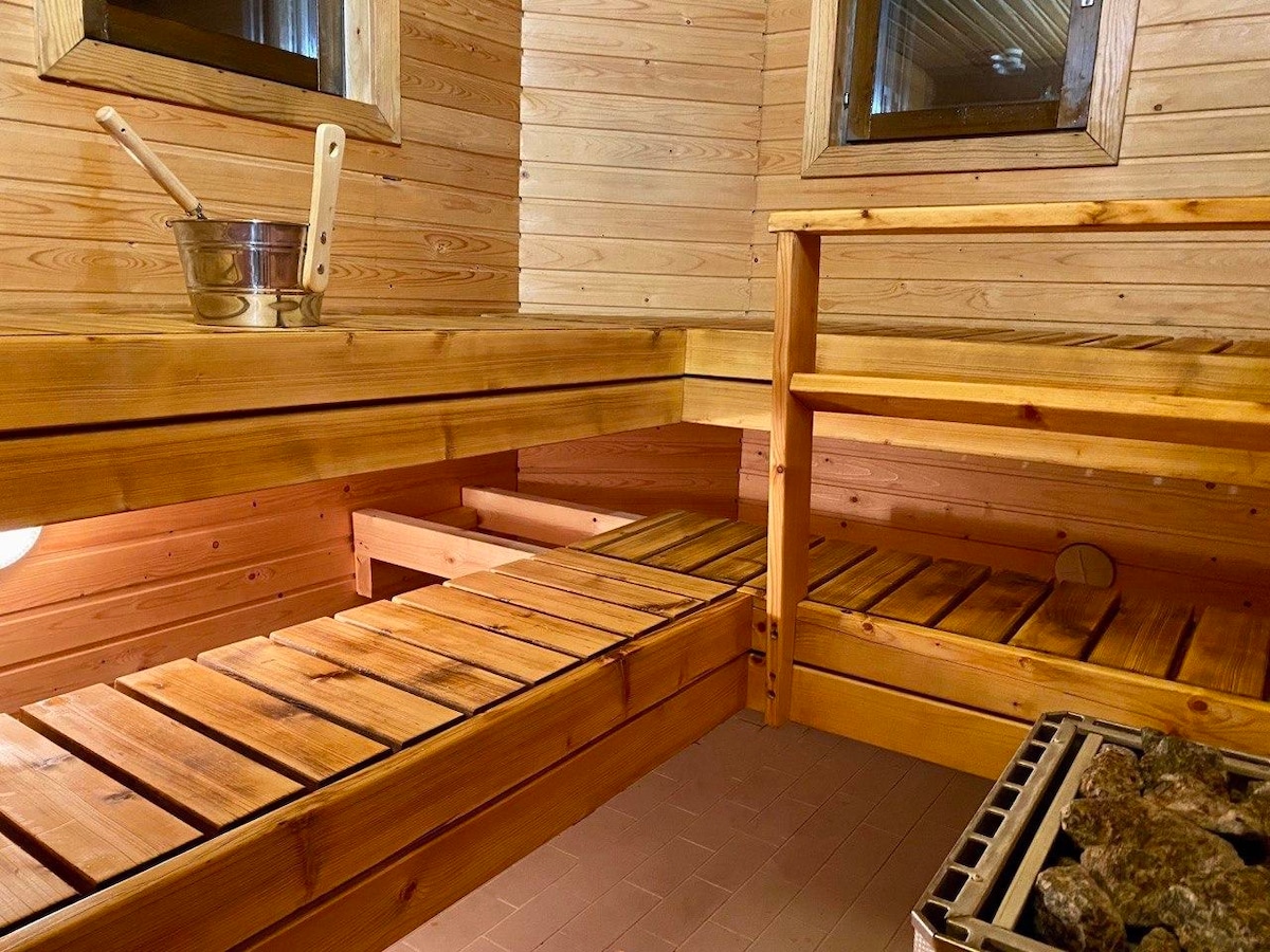 A wooden sauna interior is featured, showcasing a series of horizontal benches made from natural wood. Small windows allow light to filter in, while a stone heater is positioned in the corner, providing warmth. The setting conveys a serene atmosphere perfect for relaxation.