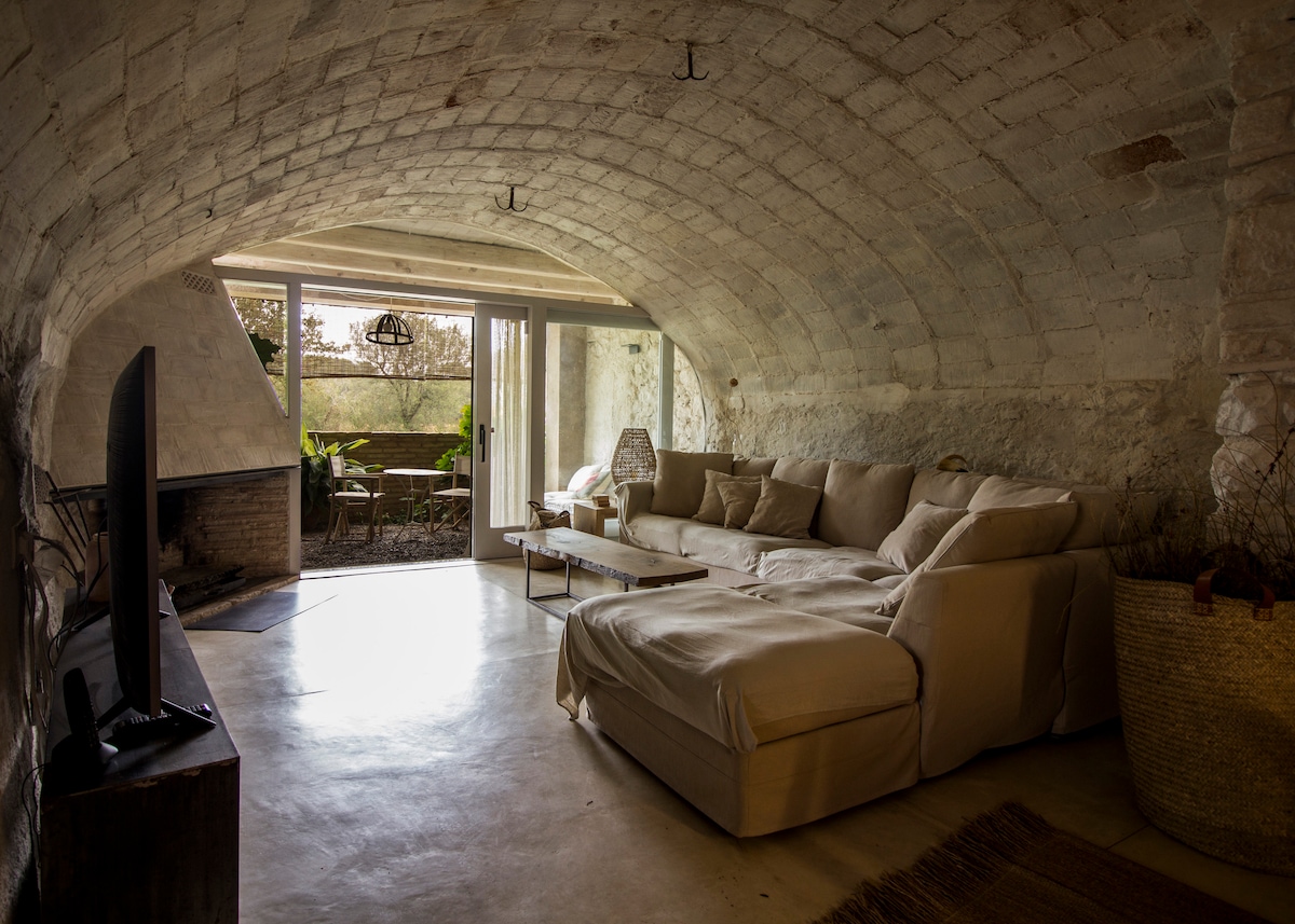 A spacious living area features a large, L-shaped sofa that invites relaxation, positioned near a flat-screen television. Natural light filters through expansive glass doors that open to an outdoor patio, enhancing the connection with nature. The room showcases rustic stone walls and a minimalist design.