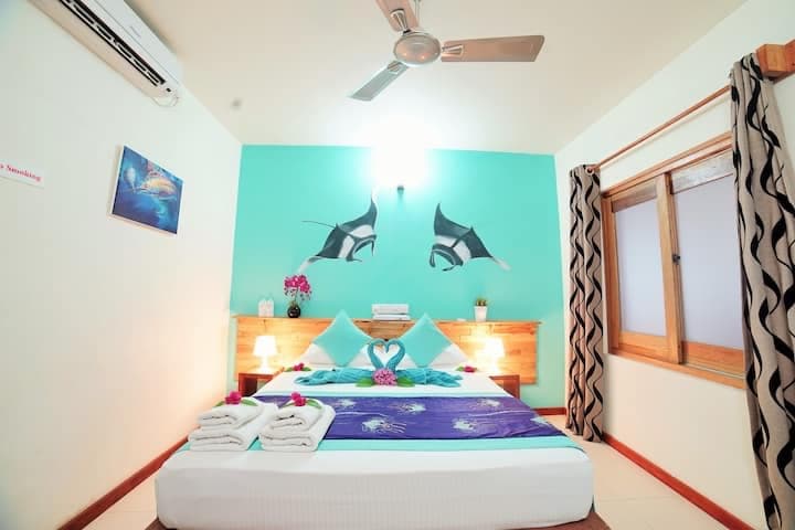 A brightly colored room features a spacious bed adorned with blue and white linen. Manta ray artwork decorates the turquoise wall above the bed. Two bedside lamps provide soft light, while a window is framed by patterned curtains, allowing natural light to fill the space.