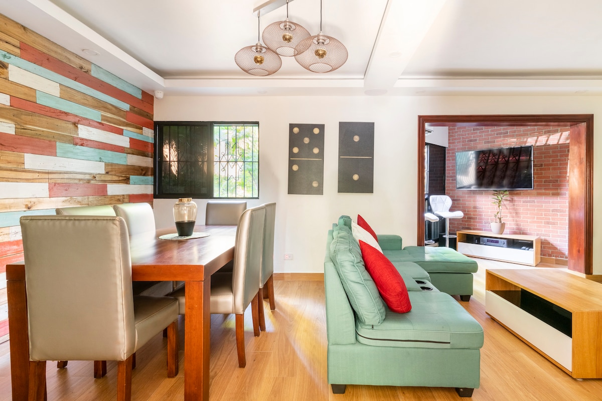 A dining area is furnished with a wooden table surrounded by six upholstered chairs. A colorful accent wall made of wooden panels is visible, along with decorative wall art. A comfortable seating arrangement includes a light green sofa with a red accent pillow. Natural light illuminates the space.