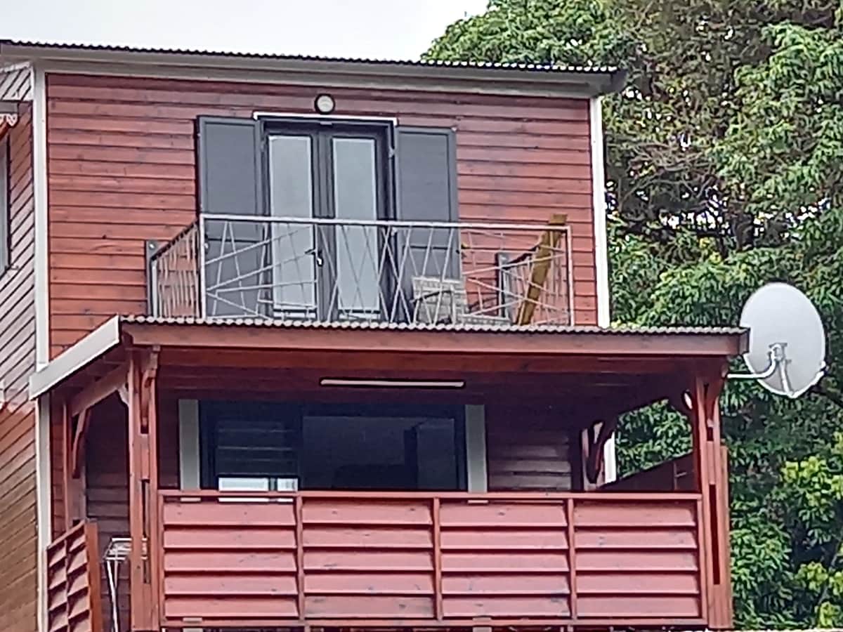 An exterior view of a two-story wooden house features a spacious balcony with a railing. Two sets of glass doors provide access to the upper floor, while a satellite dish is mounted on the side. Lush greenery surrounds the structure, enhancing its natural setting.