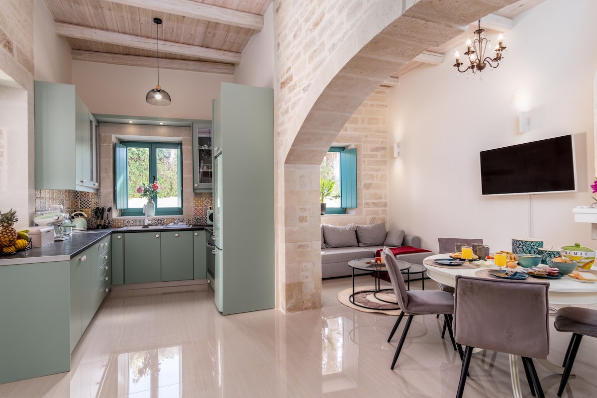A modern kitchen area with pastel green cabinets and a sleek countertop is adjacent to a cozy living space. Natural light enters through a window, illuminating a circular dining table set for breakfast, surrounded by elegant gray chairs. A comfortable sofa is positioned nearby.