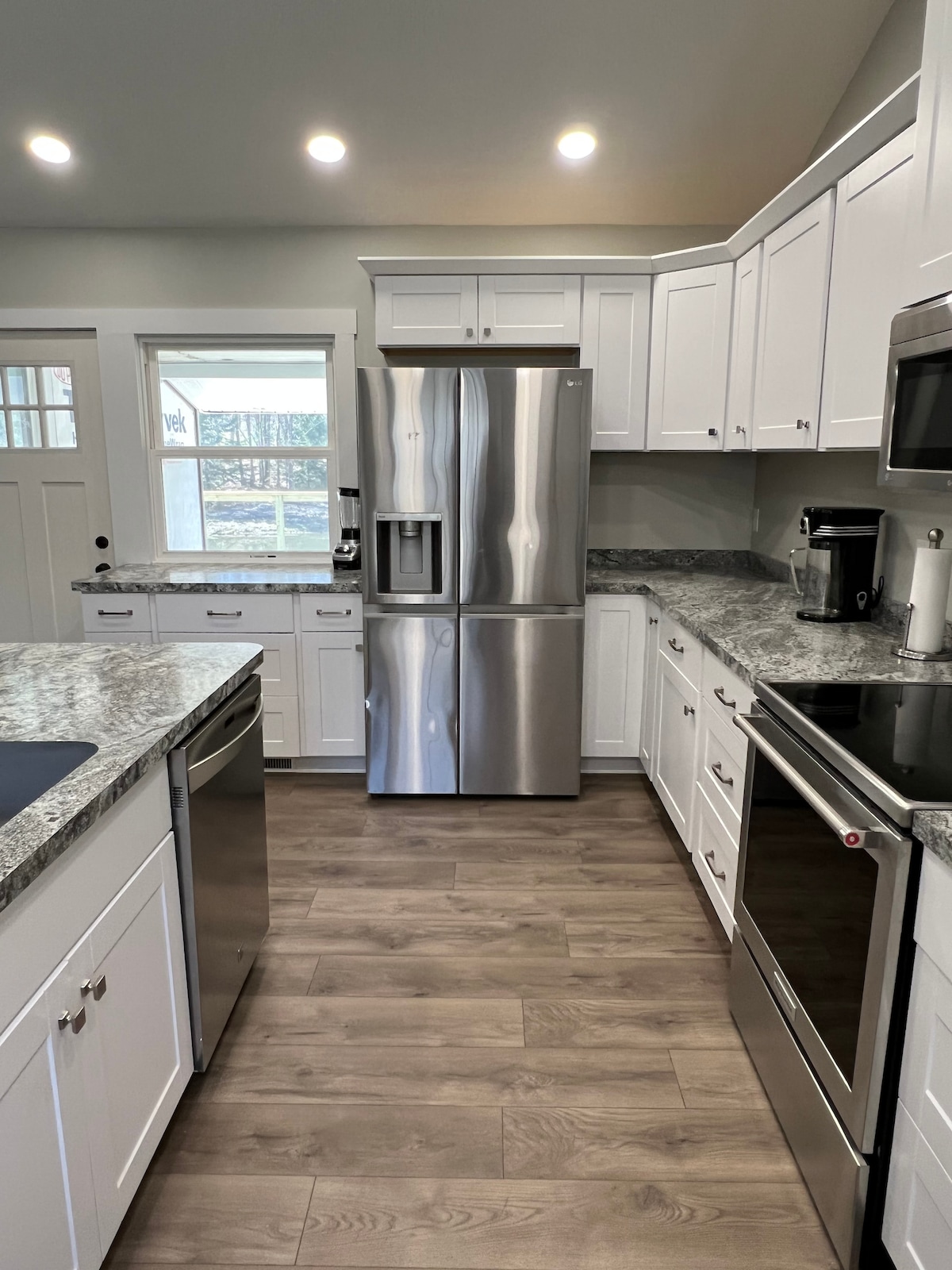 A modern kitchen is presented with white cabinetry and stainless steel appliances. A large refrigerator and a sleek oven are visible, complemented by a marble countertop. Natural light enters through a window, enhancing the welcoming and functional layout of the space.