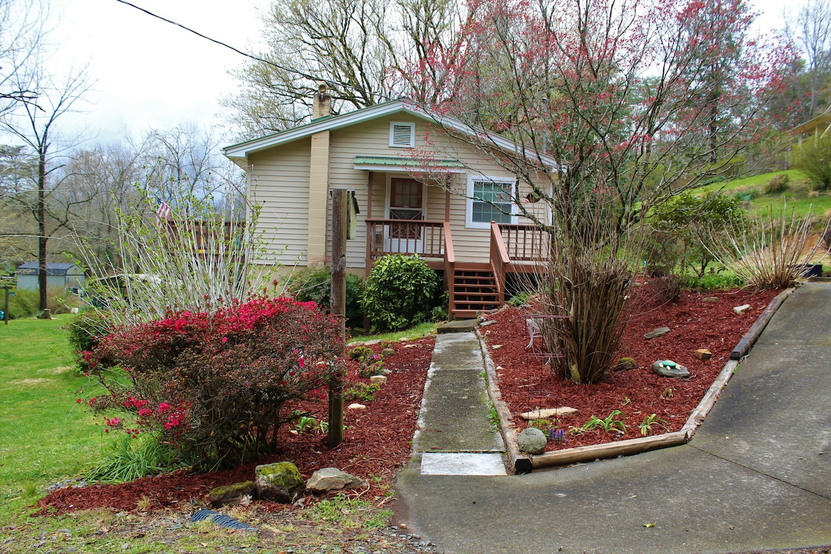 A charming exterior of the house is surrounded by well-maintained landscaping featuring flowering shrubs and ground cover. A stone path leads to the front steps, enhancing the inviting entrance. Trees frame the property, adding a sense of privacy and tranquility.