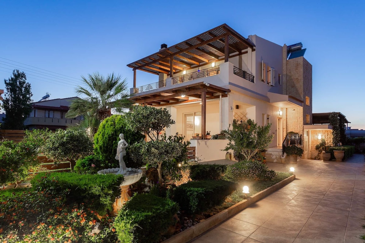 The villa's elegant exterior is framed by manicured gardens featuring lush greenery, decorative plants, and a statue. A spacious balcony with wooden beams overlooks the surroundings. Soft lighting enhances the ambience as twilight falls over the property.