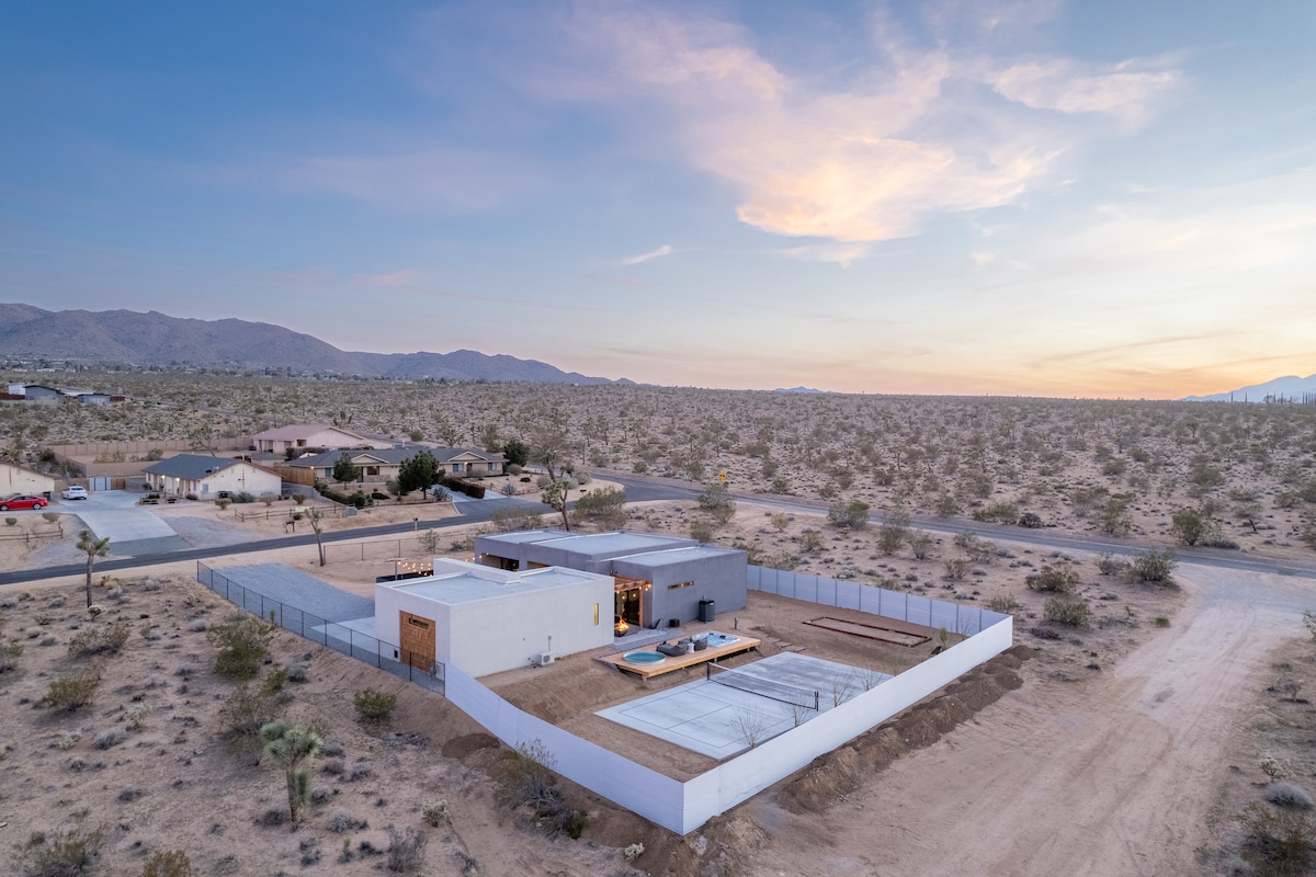 Aerial view of a modern villa surrounded by desert landscape. The spacious backyard includes a swimming pool, pickleball court, and fire pit, framed by a tall fence for privacy. Rolling hills and soft twilight colors create a serene backdrop.