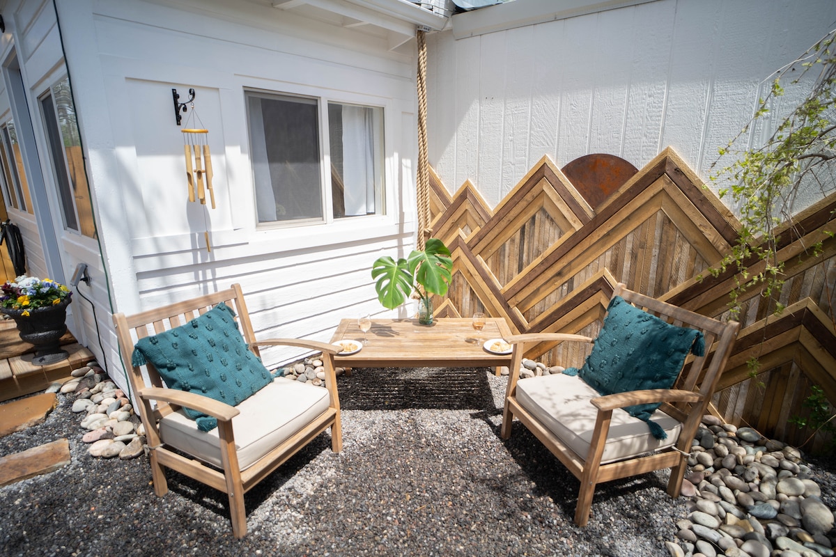 An outdoor seating area is presented with two wooden chairs featuring cushions, complemented by a wooden coffee table. Decorative elements include a small potted plant on the table and textured wooden panels in the background, creating an inviting and tranquil atmosphere.