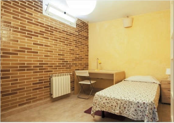 A private room features a single bed with a patterned bedspread, a desk with a lamp, and a nearby chair. A radiator is visible along the wall. The warm yellow and brick walls add texture to the space, enhancing the overall ambiance.