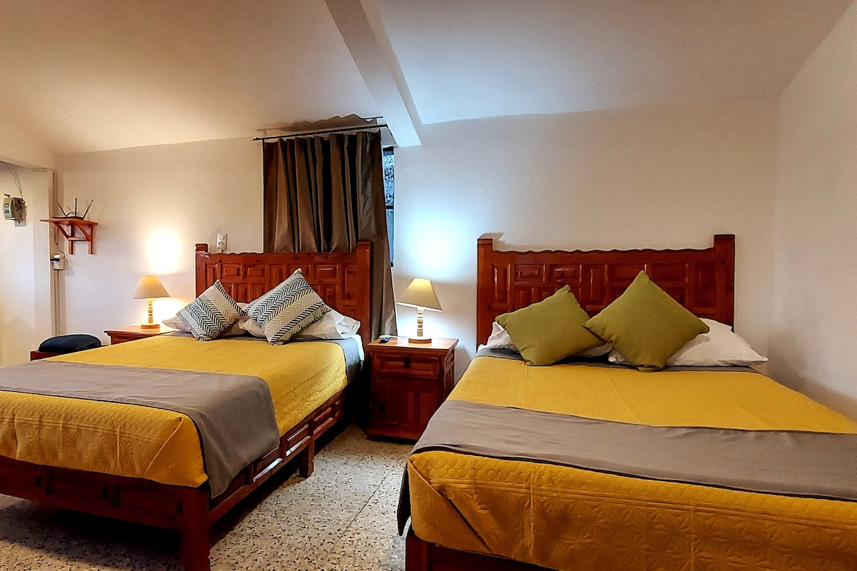 The room features two spacious beds adorned with yellow covers and decorative cushions. Nightstands with lamps are positioned beside each bed, providing soft lighting. A window with light filtering through curtains enhances the overall brightness of the space.