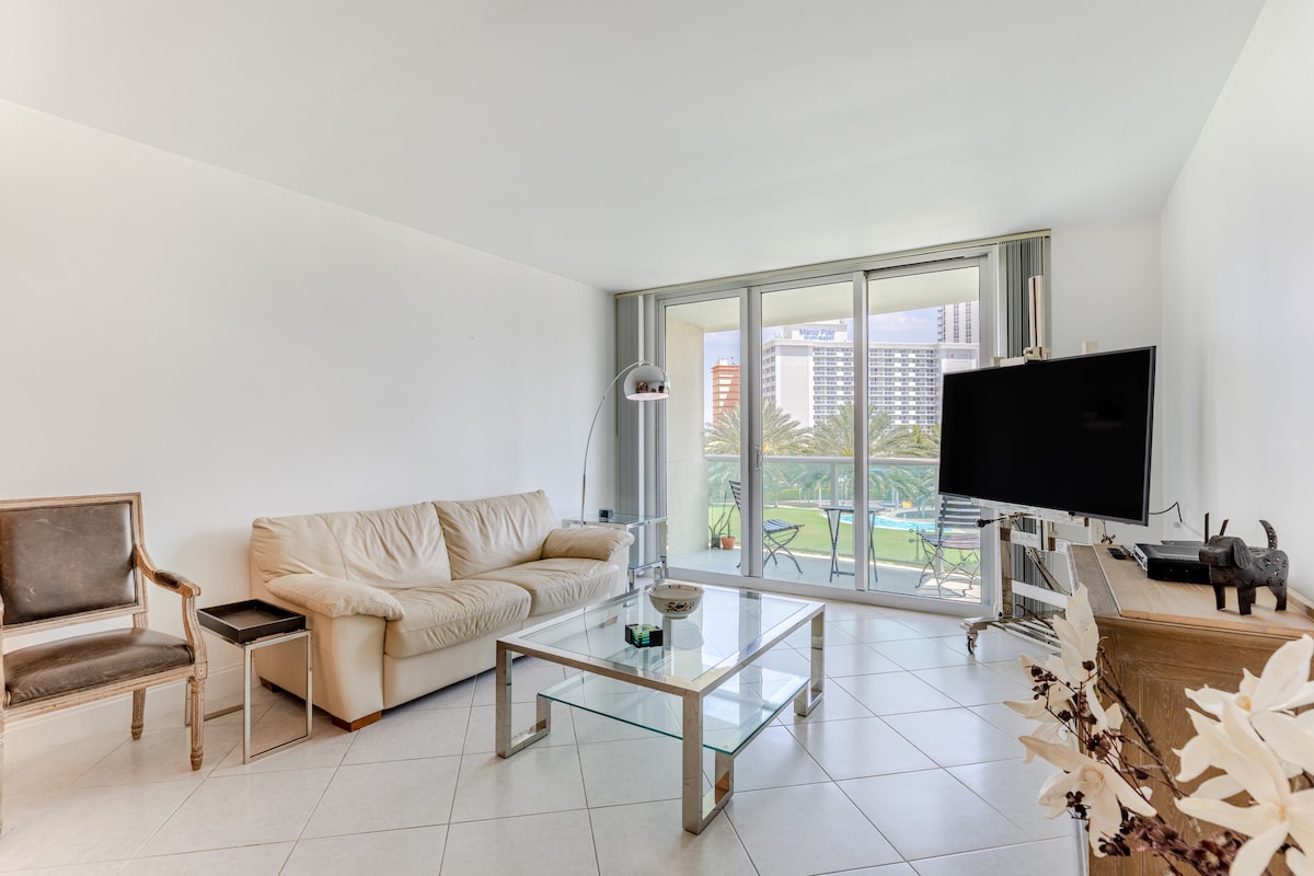 A spacious living room is showcased with a light-colored leather sofa and a glass coffee table. Large sliding doors offer views of the balcony and greenery outside. A flat-screen TV is positioned on a console, with a decorative piece displayed nearby.