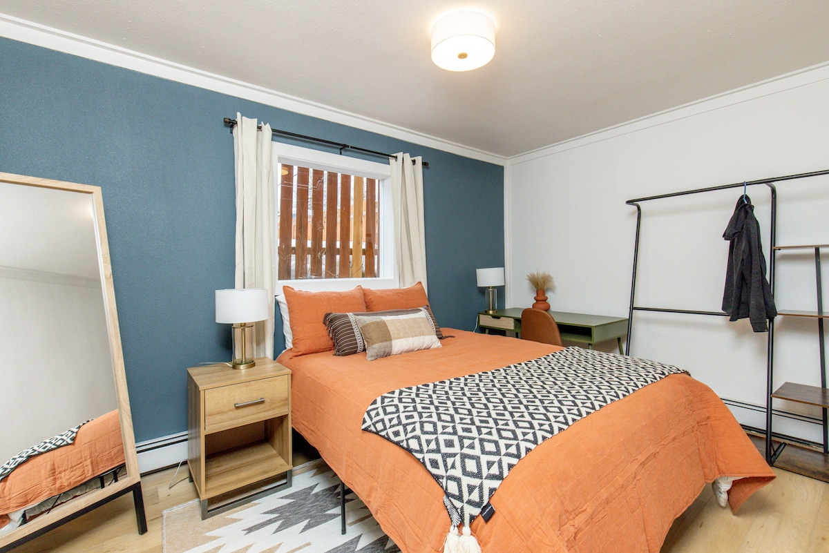 A sleeping area features a spacious bed dressed in warm orange bedding, complemented by patterned pillows. A large mirror stands against one wall, while a small desk and chair are positioned near the window. Soft lighting is provided by two bedside lamps.