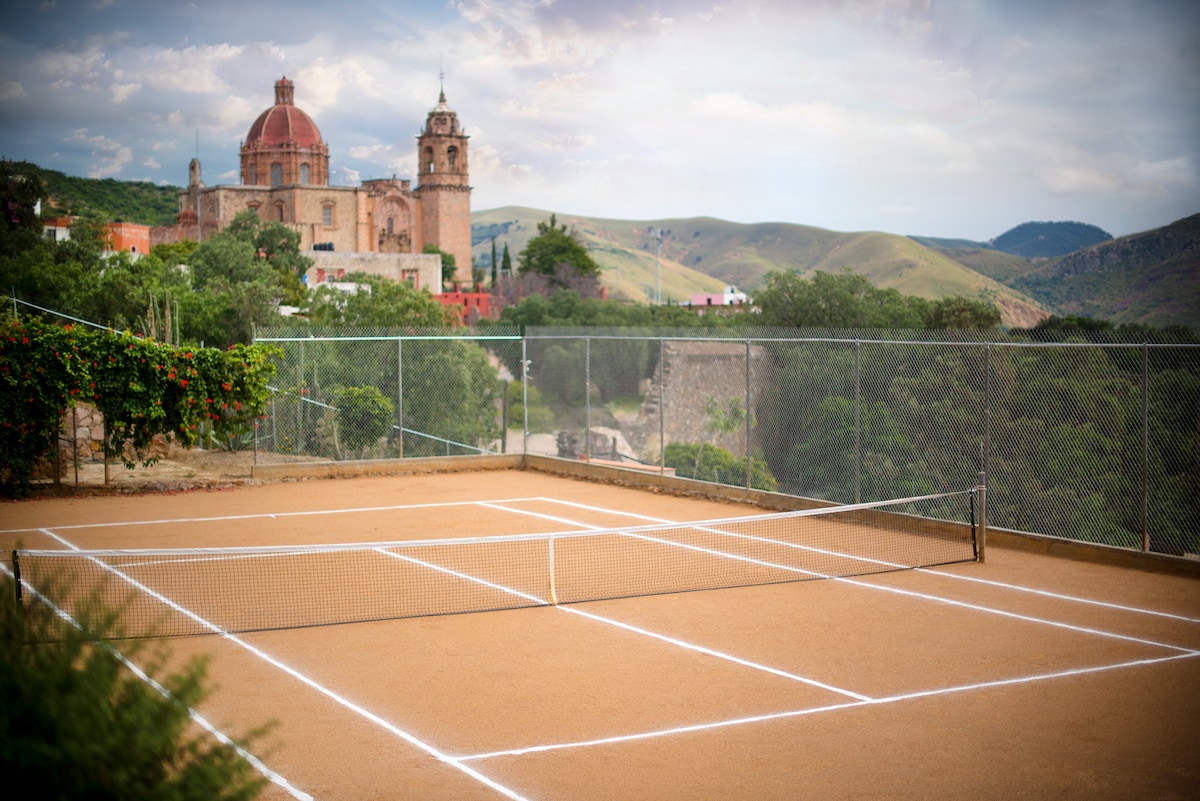 A tennis court is positioned with a view of rolling hills and a historic church in the background. The court is surrounded by a fence and greenery, highlighting a peaceful outdoor setting.