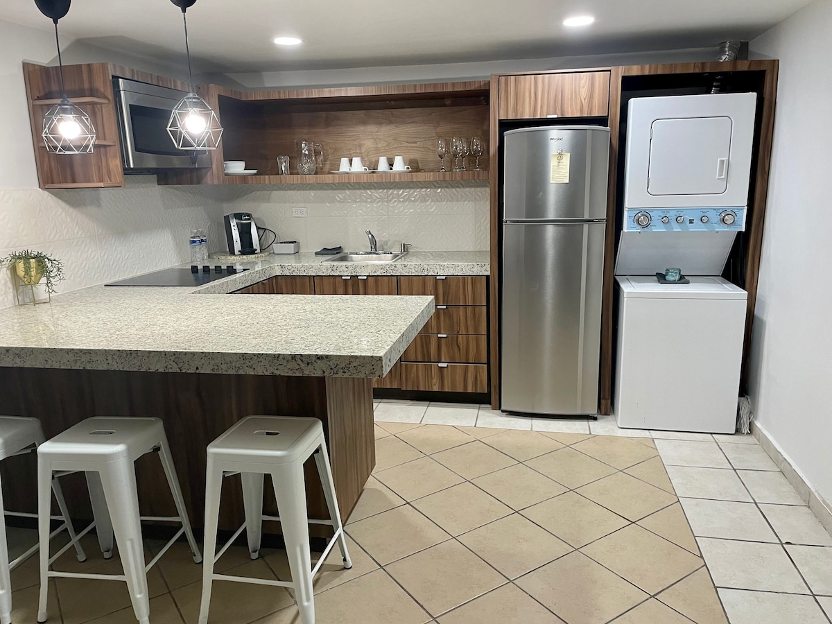 A modern kitchen features wooden cabinetry and a spacious granite countertop. Appliances include a stainless steel refrigerator, built-in microwave, and a coffee maker. A stacked washer and dryer are positioned adjacent to the kitchen, while barstools provide seating at the counter.