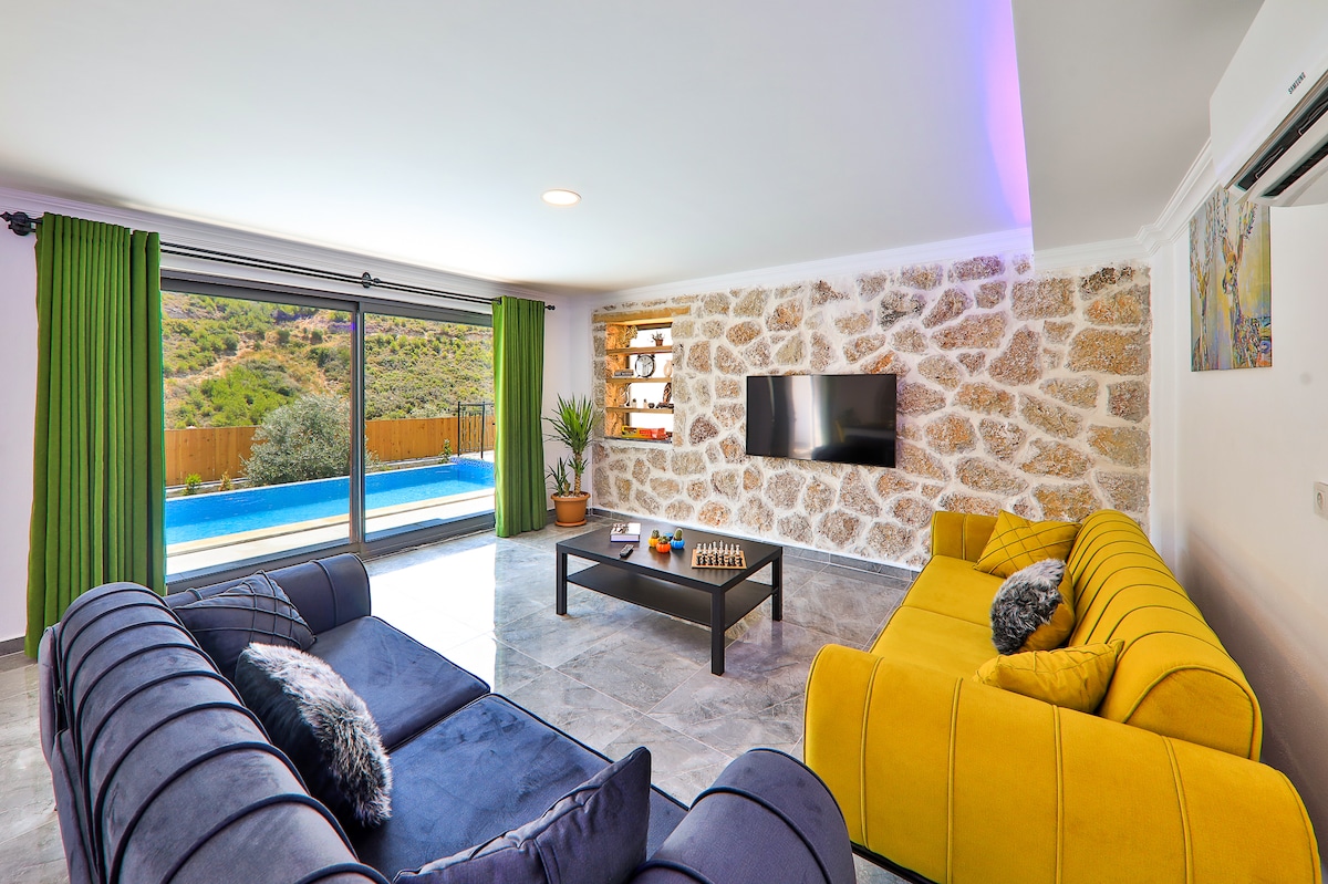 A spacious living room features a large glass door leading to an outdoor terrace with a pool view. Comfortable seating arrangements are provided by a dark blue sofa and a vibrant yellow sofa, complemented by a coffee table. Natural light fills the room, highlighting the stone wall details.