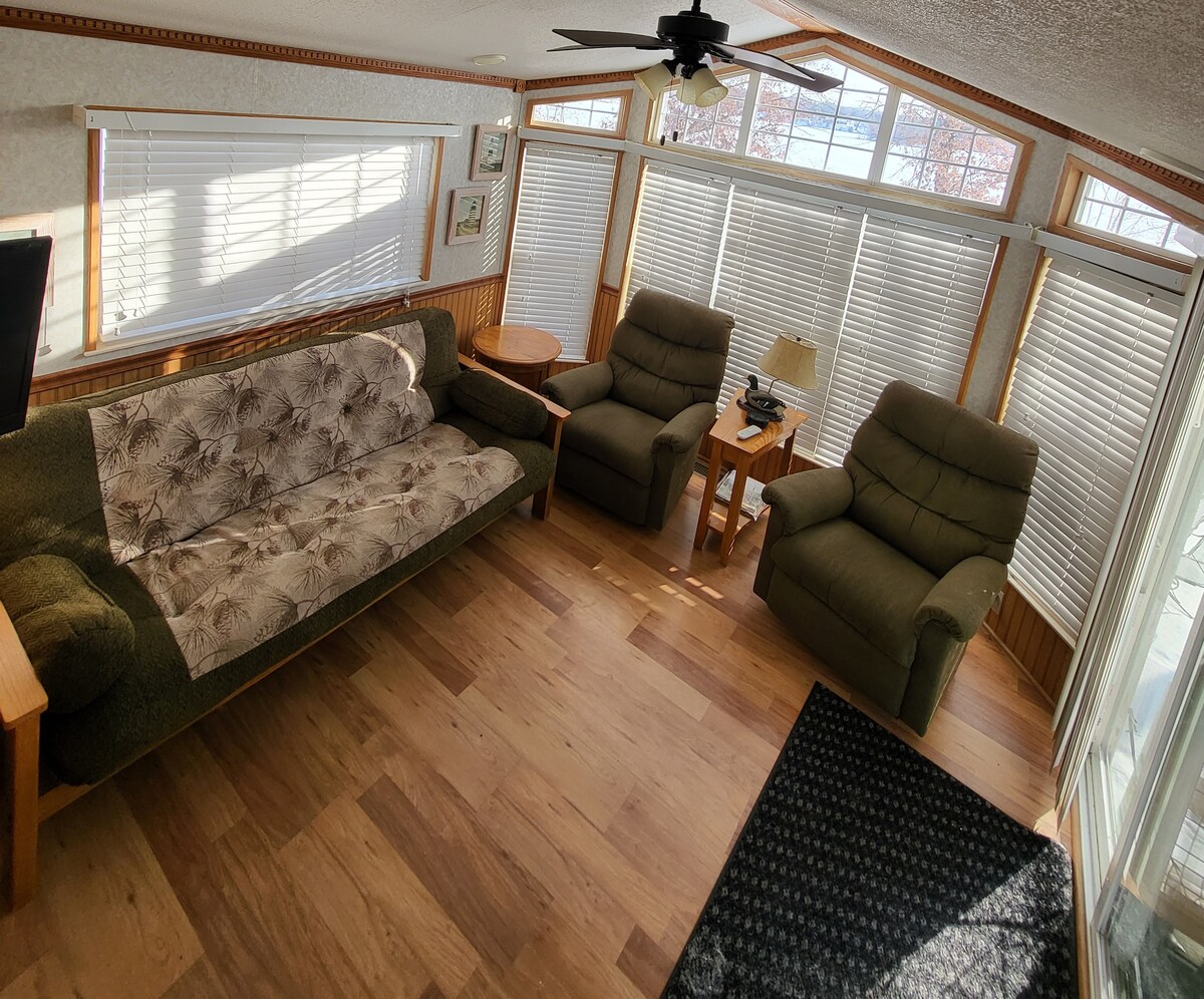 A living area features a comfortable pullout sofa and two plush armchairs arranged around a small wood table. Natural light fills the space through several windows, while light-colored walls and wooden accents create a welcoming atmosphere. A ceiling fan is positioned centrally for added comfort.