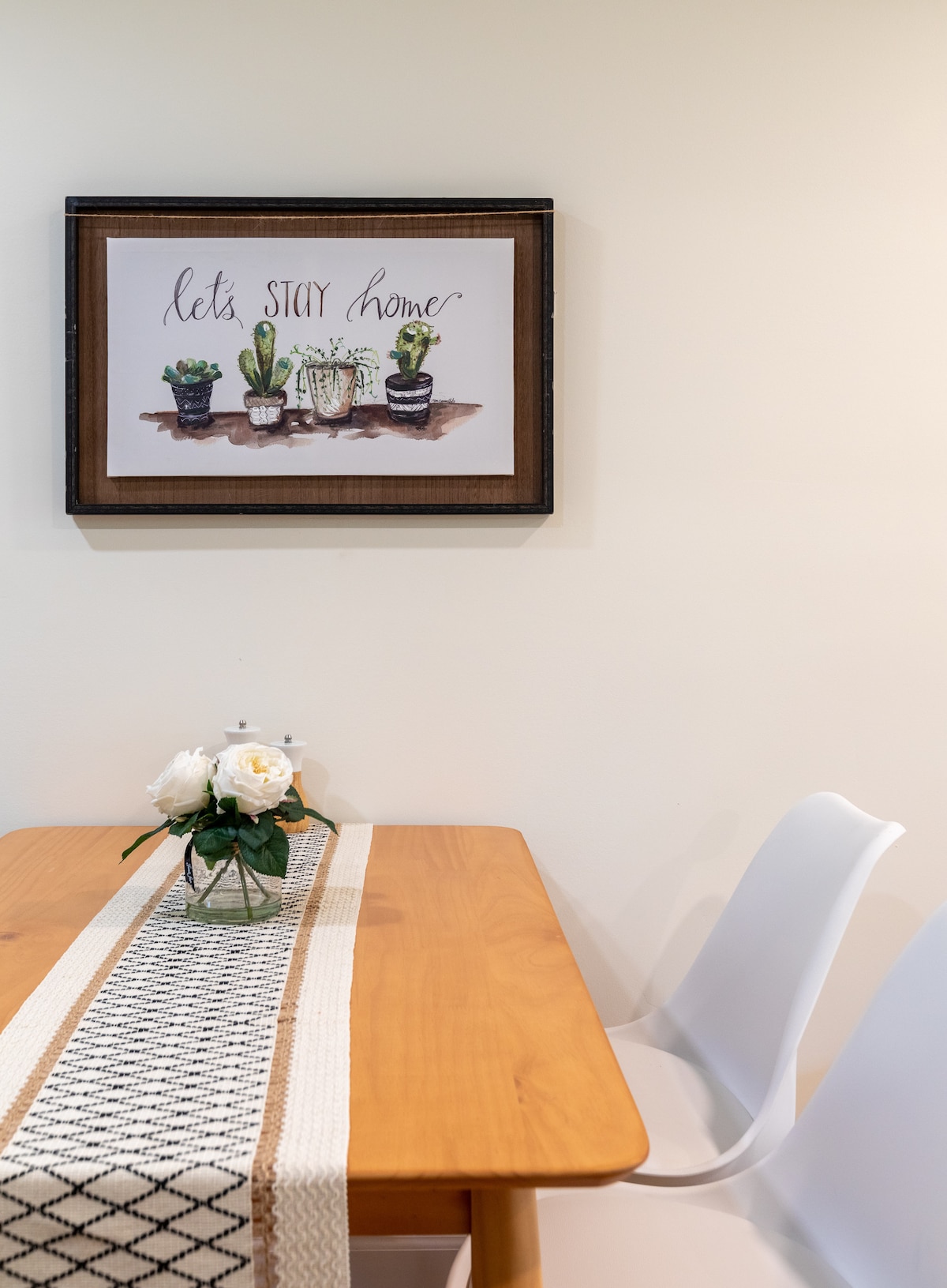 A wooden dining table is set with a neutral runner and a vase of white flowers. Two modern chairs flank the table. Above, a framed artwork featuring plants and the text 'let's STAY home' adds a welcoming touch to the dining area.