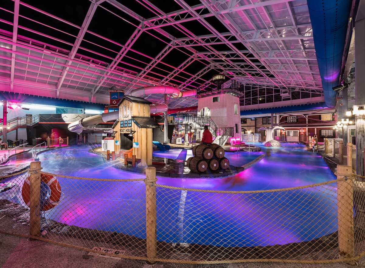 An indoor water park scene is illuminated in pink and blue lights, featuring water slides and various play structures. The pool area includes gentle waves and playful water features, surrounded by a safety fence. Seating is available near the water for relaxation.
