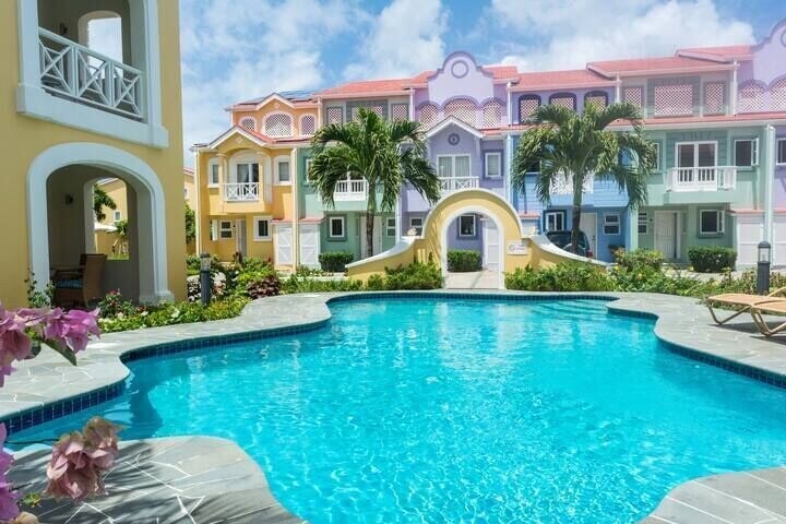 A clear, inviting pool is surrounded by lush landscaping and colorful, multi-story buildings. Sunloungers are positioned beside the pool, enhancing the tranquil atmosphere. Palm trees provide shade, and vibrant flowers add touches of color to the serene setting.