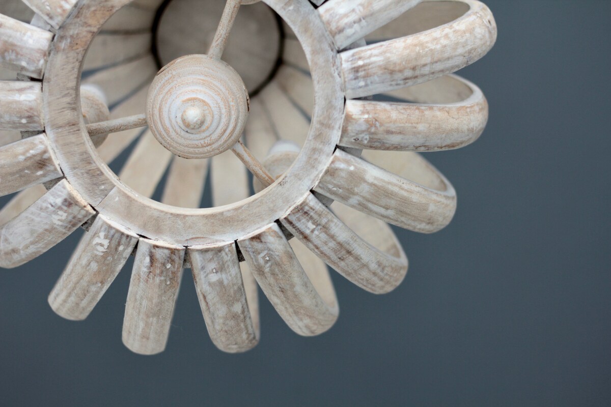 A circular light fixture features a rustic design, showcasing wooden spokes arranged in a fan-like pattern. The fixture is suspended from a ceiling, casting a warm glow on the surrounding space. A dark wall serves as a contrasting backdrop.