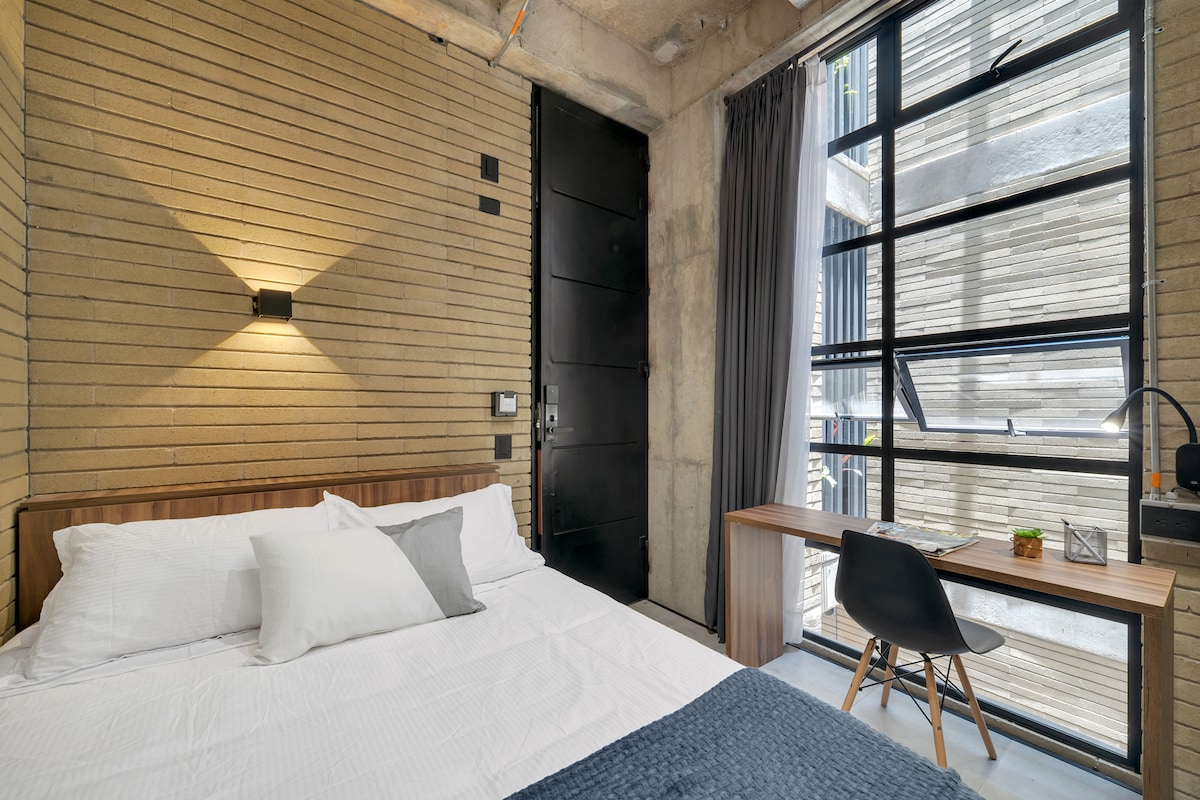 A cozy sleeping area is presented with a double bed dressed in soft white linens and a gray accent pillow. A wooden desk with a stylish lamp is positioned beside a large window, allowing natural light to fill the space. Textured, exposed brick walls add a unique touch.