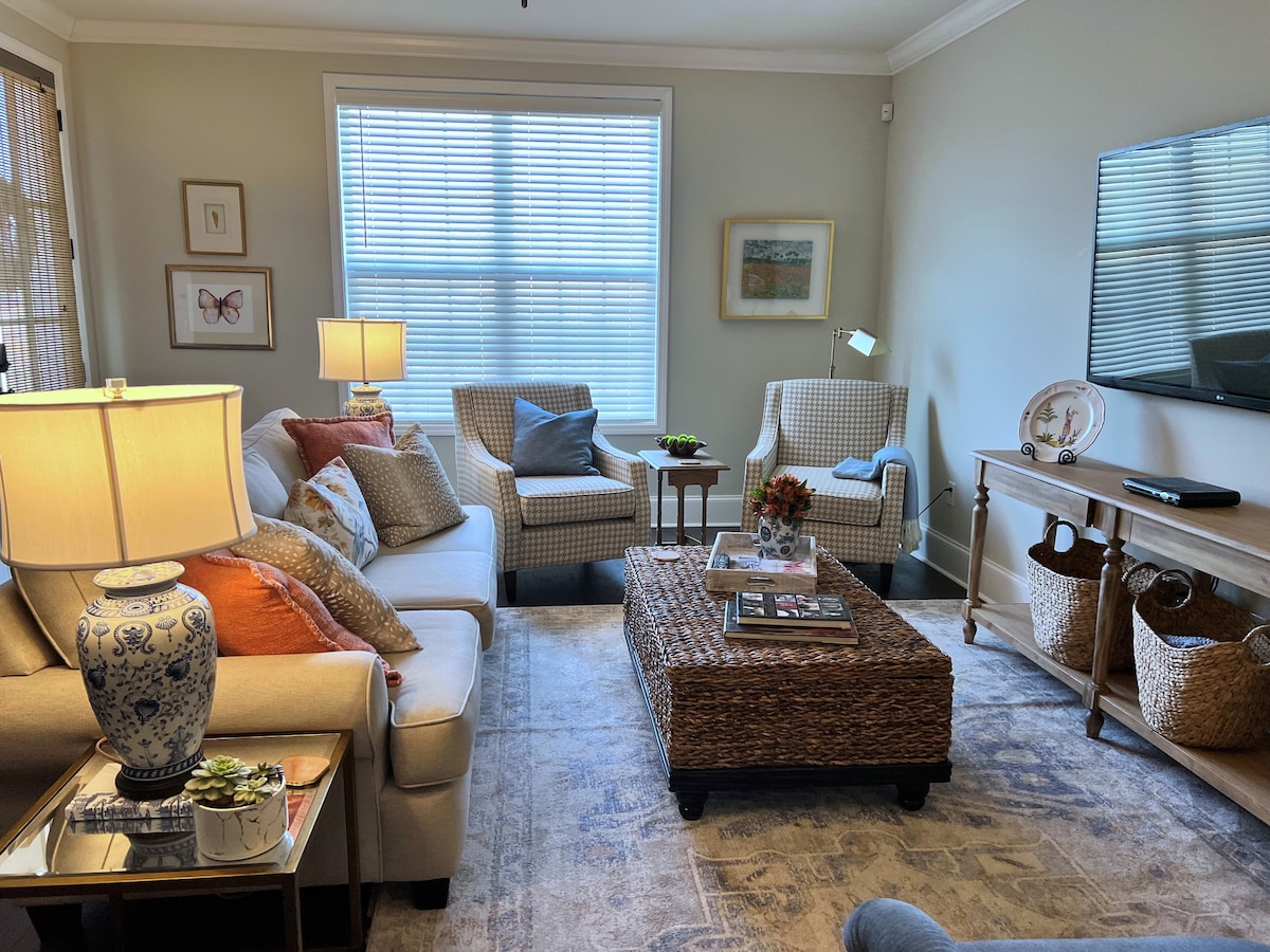 A welcoming living area features a soft sectional couch accented with colorful throw pillows, flanked by two armchairs. A coffee table made of natural materials stands at the center, while soft light filters through the window, illuminating the space.