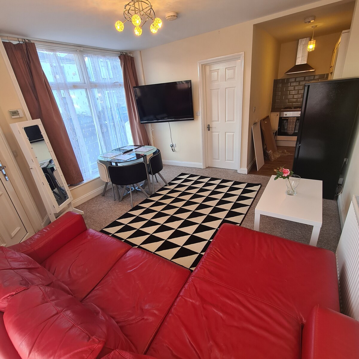 A spacious living area showcases a red corner sofa and a modern black and white patterned rug. A glass dining table with chairs is positioned near large bay windows, which provide ample natural light. A wall-mounted television is visible, along with a glimpse of the kitchen area.