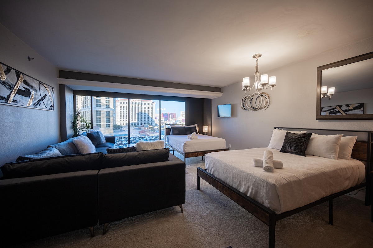 A spacious suite features two king beds and an L-shaped couch, complemented by large windows that offer a view of the city skyline. Elegant lighting is provided by a chandelier, and a framed mirror enhances the ambiance.