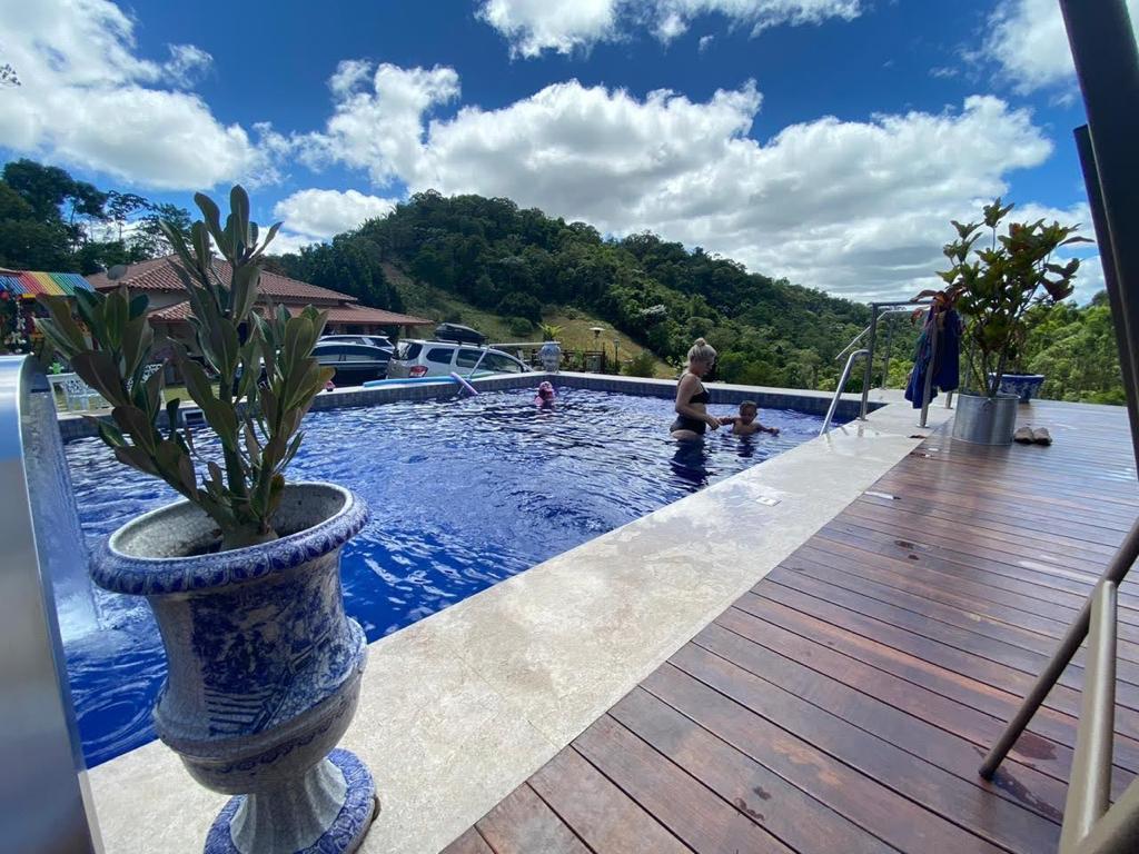 A large swimming pool is surrounded by a wooden deck, with steps leading into the water. Potted plants are placed along the edge. Guests are seen enjoying the water, with green hills and a cloudy sky visible in the background.