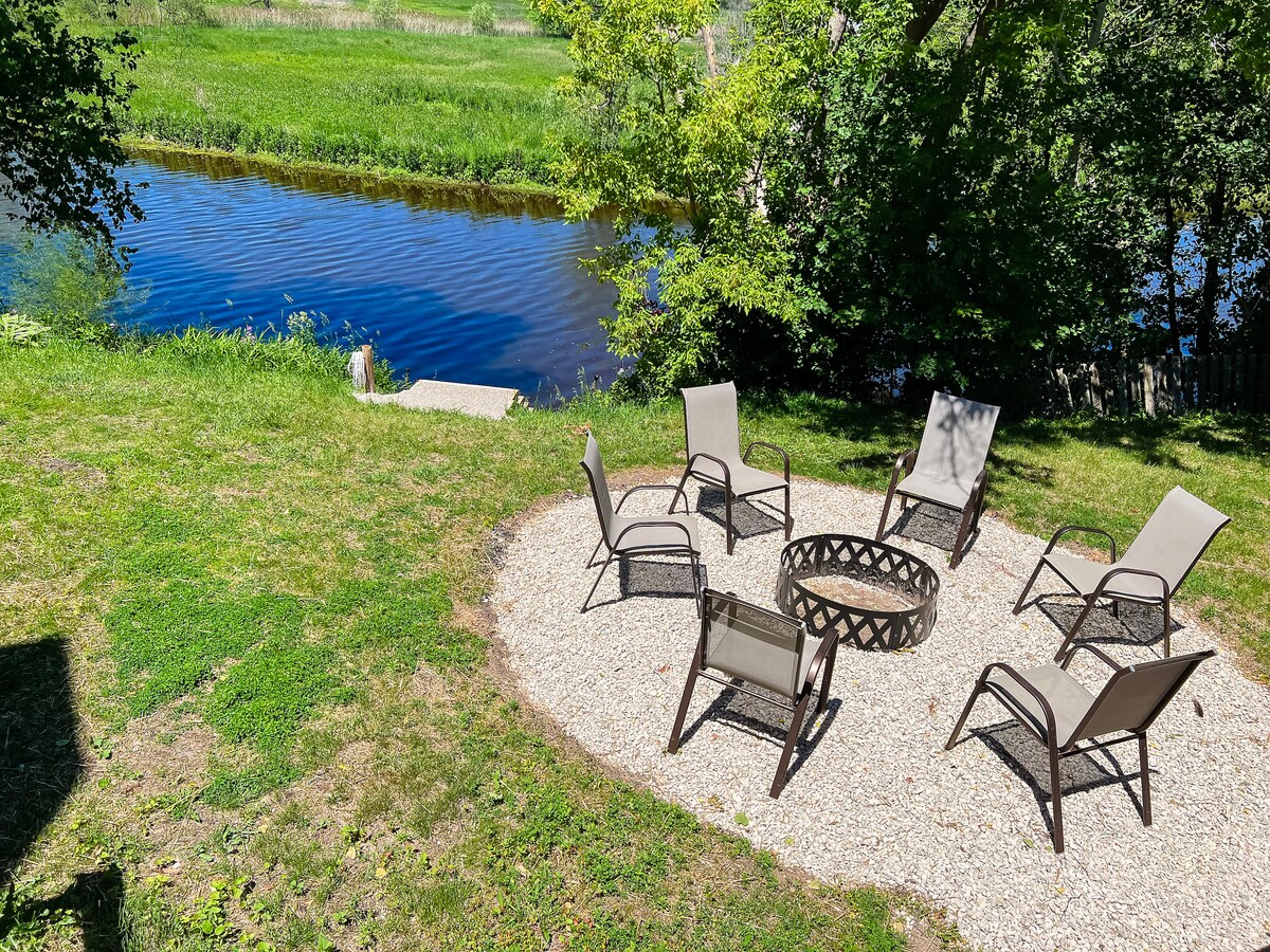 A gravel seating area is arranged with six comfortable chairs around a central fire pit, set against a backdrop of lush greenery and a serene river. The tranquil water reflects the surrounding landscape, offering a peaceful outdoor retreat.