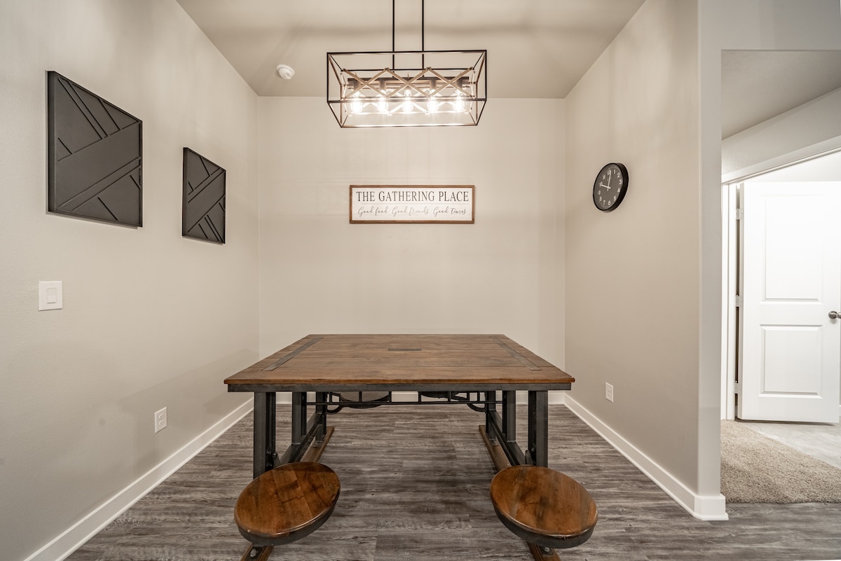 A dining area is featured with a large wooden table and benches, set against a light wall. Above, a modern light fixture provides illumination. Decorative wall art and a clock add character, creating a welcoming space for meals or gatherings.