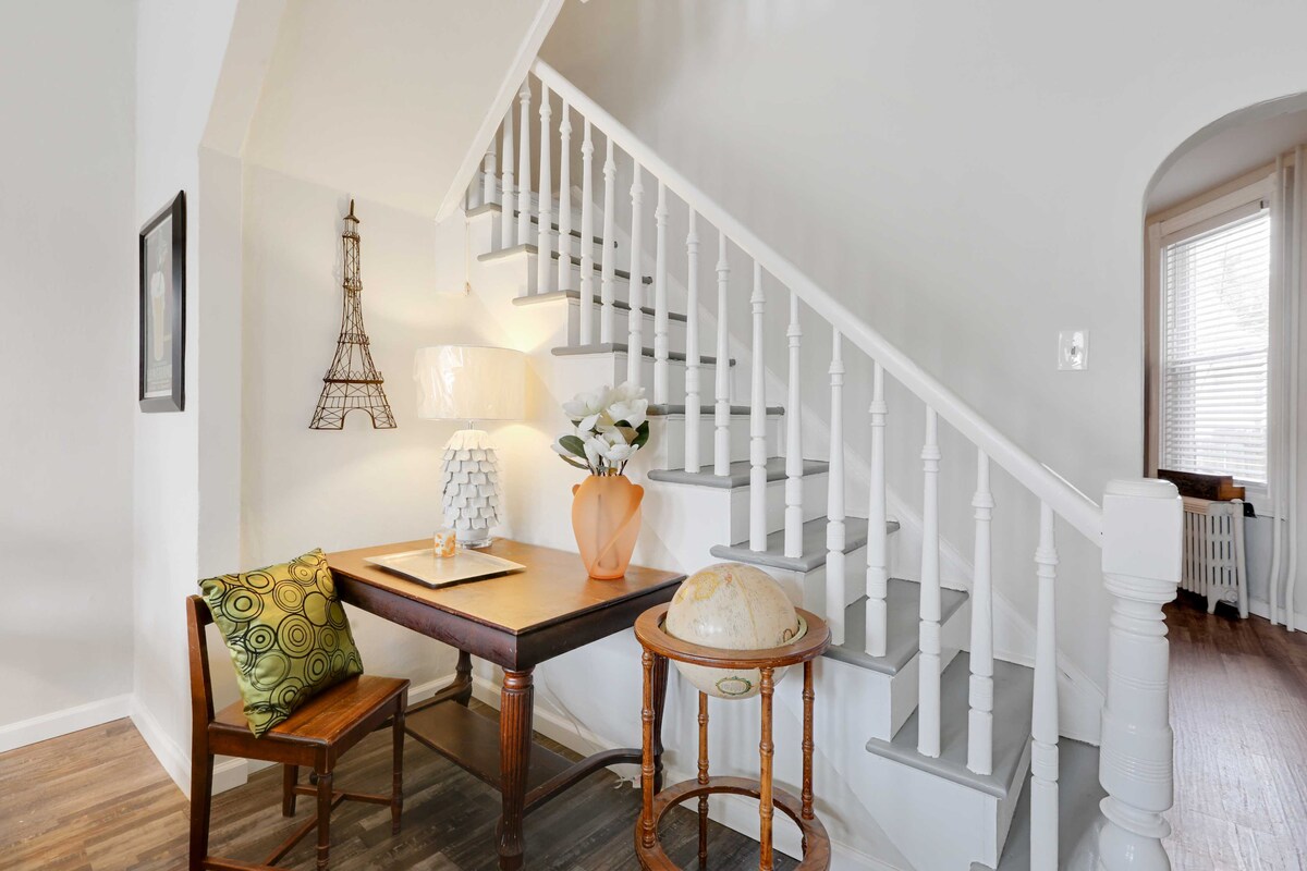 A bright entryway features a staircase with white railings and wooden steps. A small desk and chair are placed against the wall, accompanied by a decorative globe and a vase with flowers. Natural light enters through a window, illuminating the space.