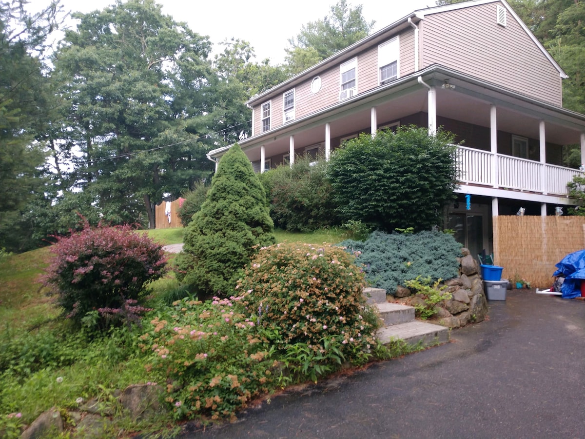 A two-story house is situated on a slightly elevated lot, surrounded by well-maintained foliage. A mix of flowering shrubs and evergreen trees frame the entrance, leading up to a welcoming porch with white railings. A paved driveway is visible, providing access to the side of the home.