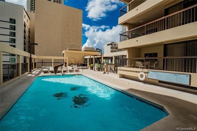 A refreshing pool area is depicted, featuring clear blue water under a bright sky. Sun loungers are arranged around the poolside, providing space for relaxation. The surrounding terrace is equipped with shaded seating areas, and the building's architecture is visible in the background.