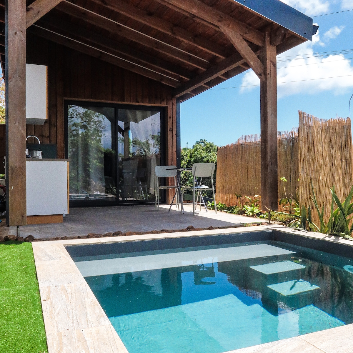 A covered patio area features a stone-tiled surface, leading to a swimming pool with steps visible on one side. A compact dining setting with two chairs is placed nearby, surrounded by greenery. Large glass doors open to an indoor space, enhancing the connection with nature.