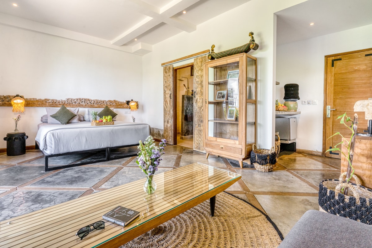 The spacious apartment features a comfortable bed with neutral bedding, surrounded by natural wood accents. A coffee table with a magazine is positioned on a woven rug. Soft lighting is provided by wall-mounted lamps, enhancing the serene atmosphere. A kitchenette is visible in the background.
