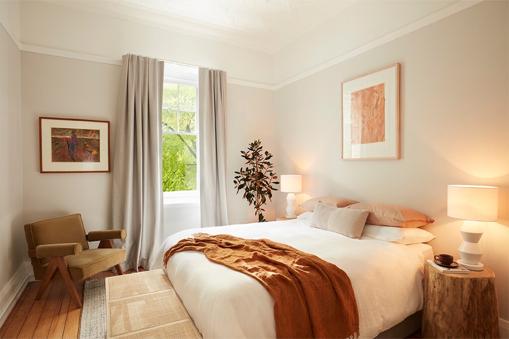 A serene bedroom setup features a spacious bed adorned with neutral linens and a warm throw. A minimalist chair complements the space alongside a small wooden side table, while natural light filters through a window draped with light curtains, offering a glimpse of greenery outside.