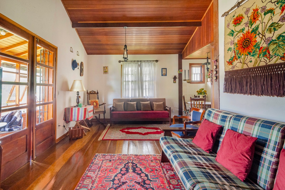 A warm living room is displayed with wooden ceilings and polished floors. A mix of seating options, including a sofa and armchair, surrounds a decorative area rug. Natural light enters through large windows, illuminating the wall adorned with colorful artwork and a vibrant textile hanging.